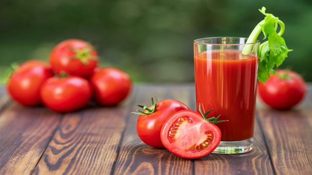 Glass of fresh tomato juice garnished with celery stick on a wooden table, surrounded by whole ripe tomatoes and a halved tomato in the foreground